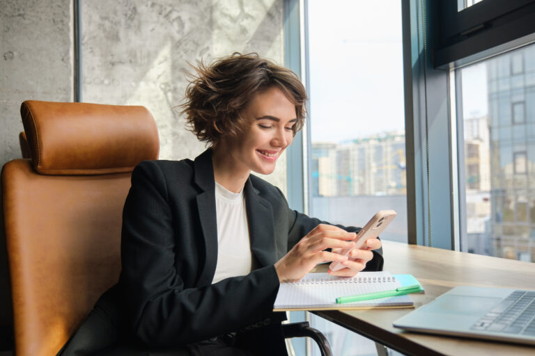 Portrait of corporate woman in suit, sits in office, messaging someone, replying to friend on smartphone, working with laptop, having lunch break.